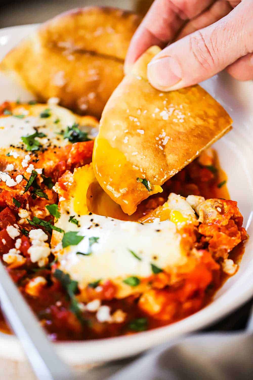A person dipping a toasted pita triangle into a poached egg nestled into shakshuka.