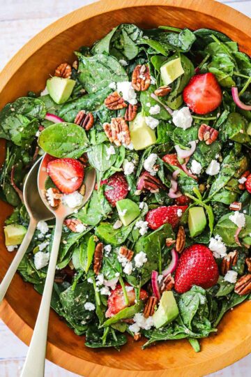 An overhead view of a fresh strawberry spinach salad with avocado that has been tossed together in a wooden bowl.