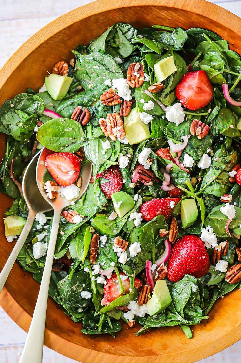 An overhead view of a fresh strawberry spinach salad with avocado that has been tossed together in a wooden bowl.