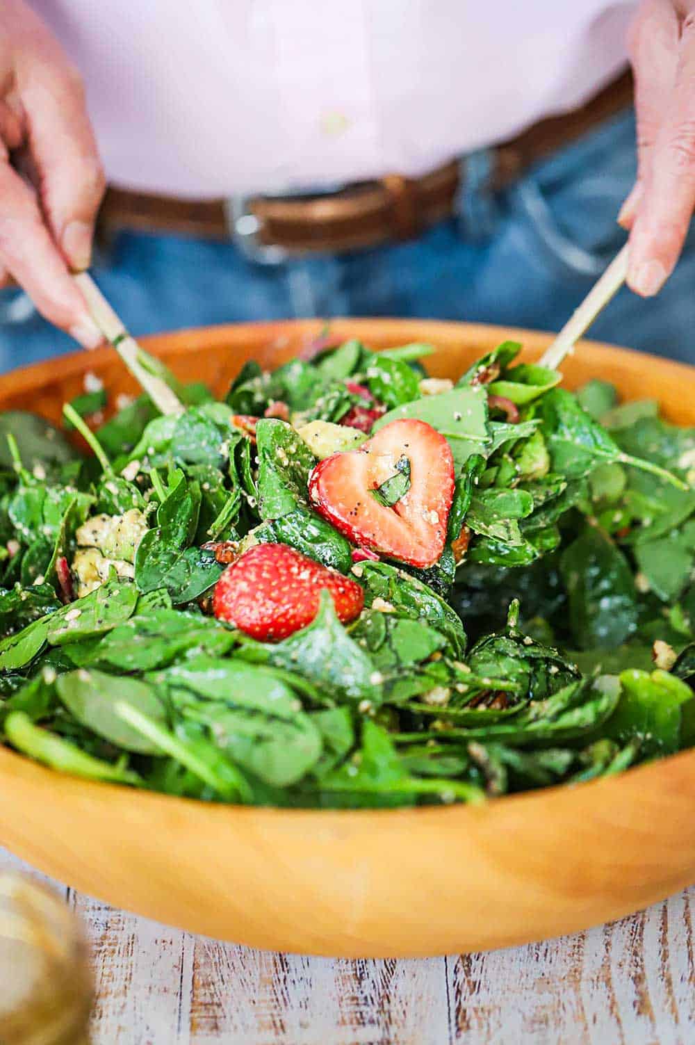 A person using two large utensils to toss a salad of fresh strawberries, spinach, and avocado with a balsamic vinaigrette in a large wooden bowl.