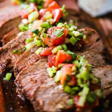 A close-up view of a grilled trip-tip with Santa Maria salsa on a cutting board next to a large knife.