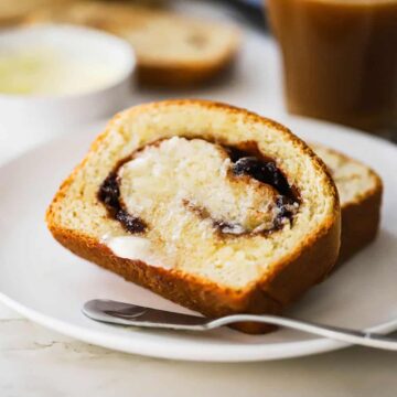 Two slices of cinnamon raisin bread stacked on top of each other with melted butter on one of the slices all on a plate next to a mug of coffee.
