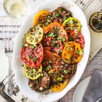 An overhead view of a large white oval platter filled with a sliced heirloom tomato salad and surrounded by a glass of white wine, olive oil, and white plate.