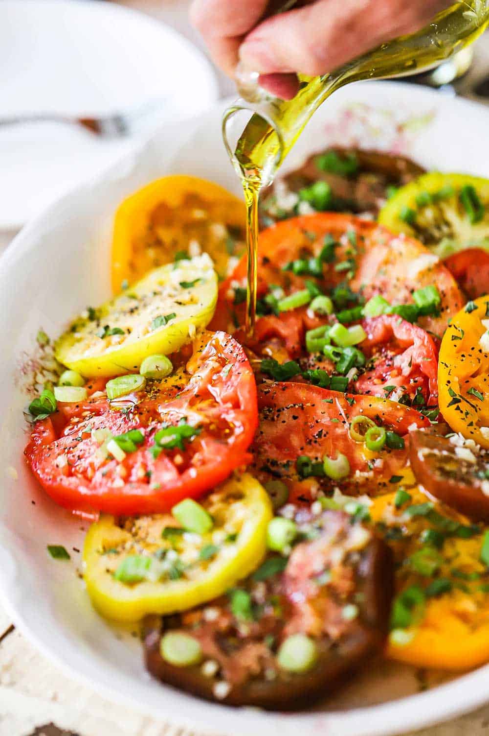 A person pouring olive oil from a glass container onto a platter filled with a heirloom tomato salad.