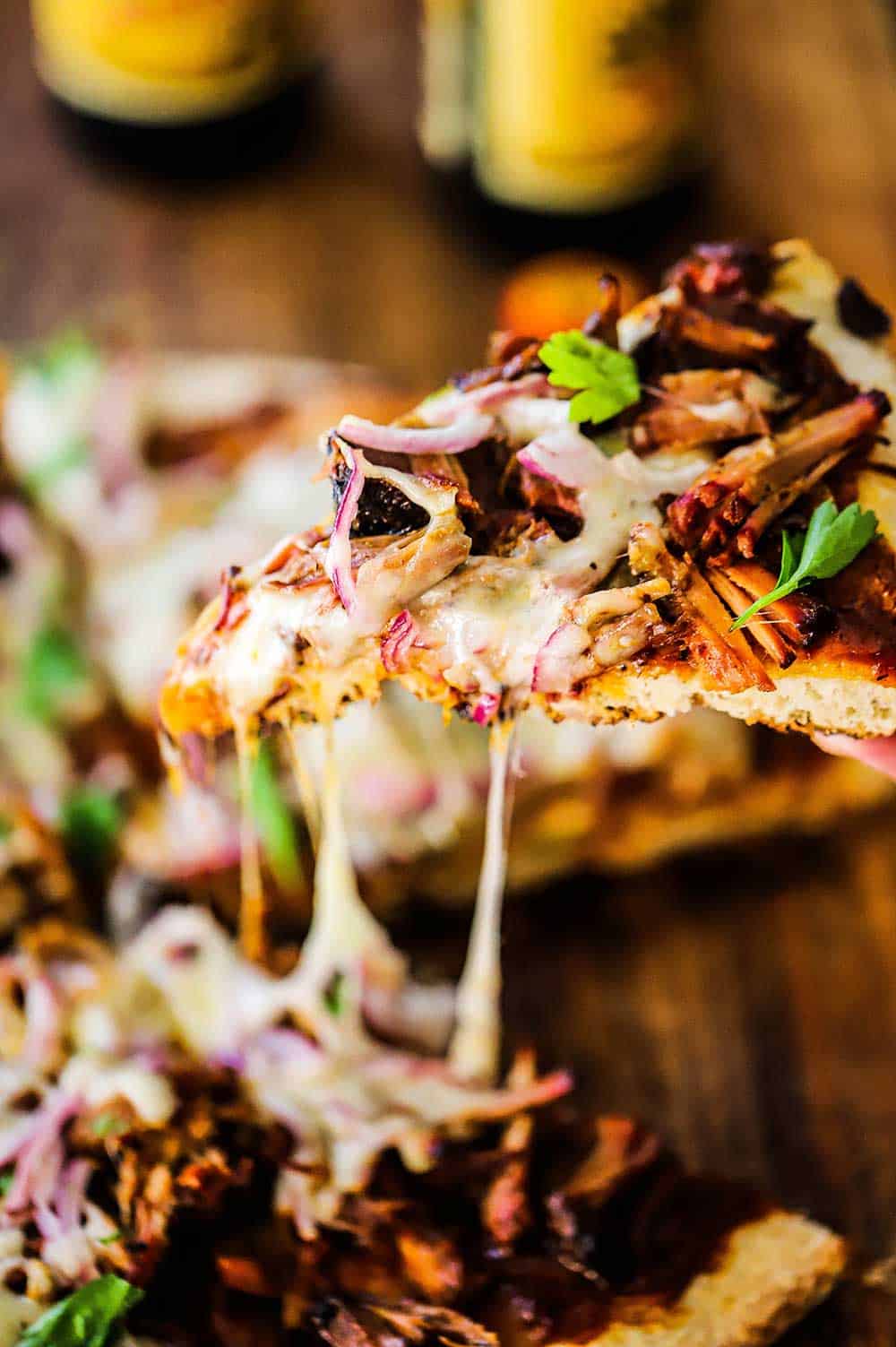 A slice of pulled pork pizza that is being raised from the whole pizza with a several strands of cheese being pulled.