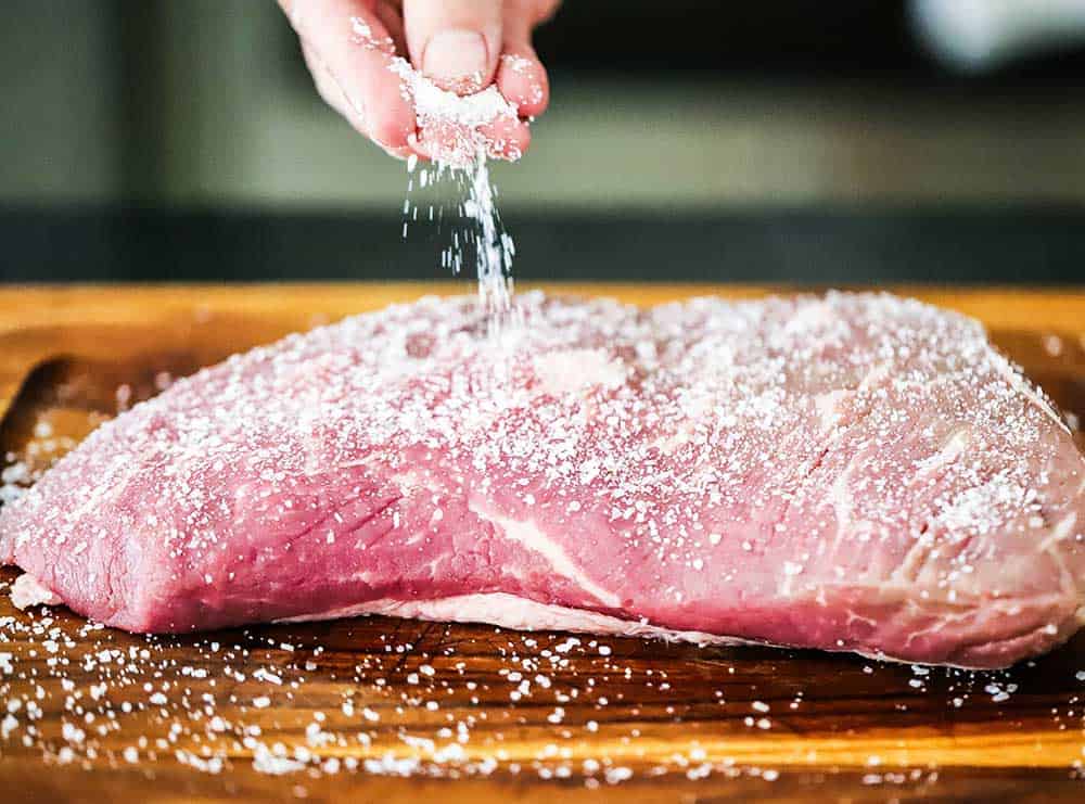 A person sprinkling coarse salt all over an uncooked tri-tip steak.