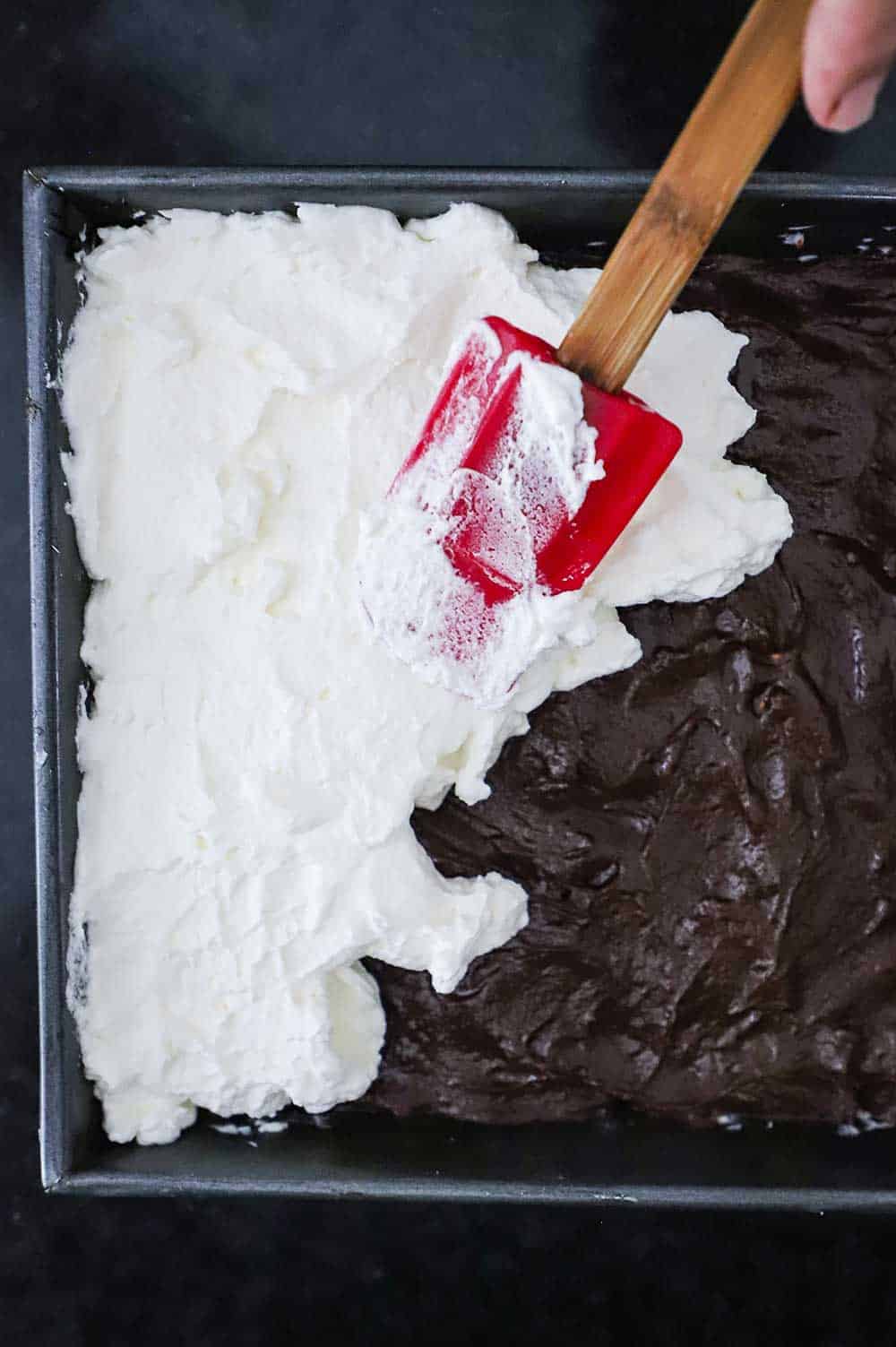 A red spatula being used to spread whipped cream topping over a layer of homemade chocolate pudding in a metal baking pan.
