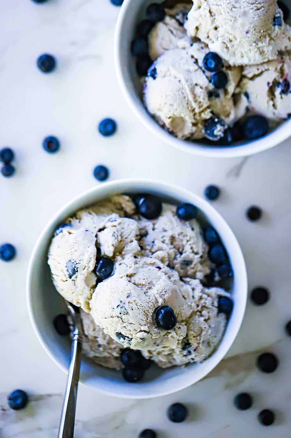 An overhead view of two ice cream bowls filled with homemade blueberry ice cream with fresh blueberries scattered all around the bowls.