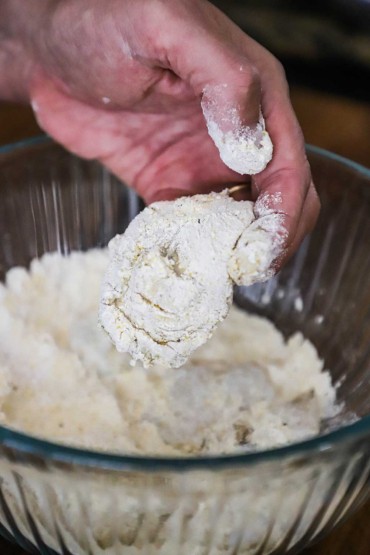 A person's hand holding a raw oyster that is covered with a flour and cornmeal mixture over a bowl of the same.