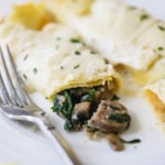 Two savory crêpes on a plate with one that has been broken open with mushrooms and spinach spilling out of it.