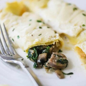 Two savory crêpes on a plate with one that has been broken open with mushrooms and spinach spilling out of it.