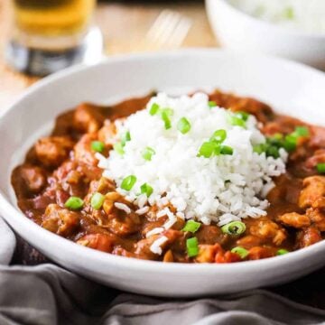 A close-up view of a white bowl filled with chicken creole with steamed white rice on top garnished with chopped scallions.