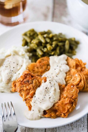 A close-up view of two Southern-fried pork chops on a dinner plate with cream gravy drizzled over the top of them and sitting next to mashed potatoes and green beans.