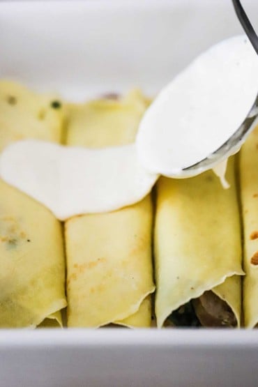 A ladle being used to transfer a creamy cheese sauce over a row of savory crêpes stuffed with sausage and mushrooms.