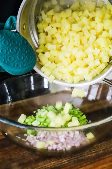 A person dumping cooked cubed potatoes into a glass bowl filled with chopped onions and celery.