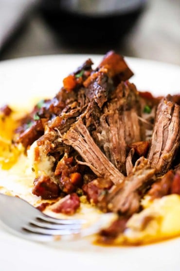 A plate filled with braised beef short ribs that have been pulled apart with a fork all on top of mashed potatoes.