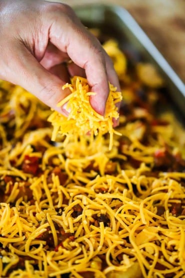 A person sprinkling finely shredded cheddar cheese over the top of a pan of nachos.