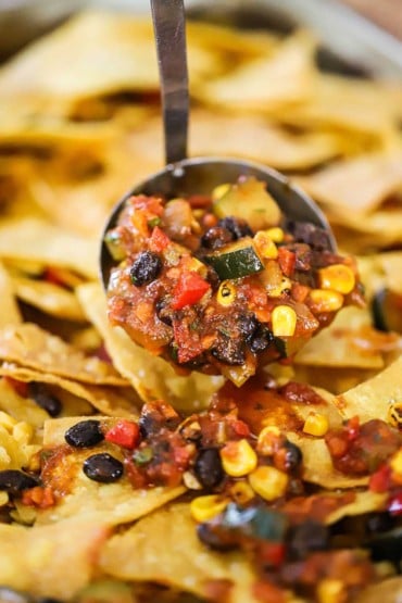 A large ladle being used to spread black bean and veggie chili over fried tortilla chips.