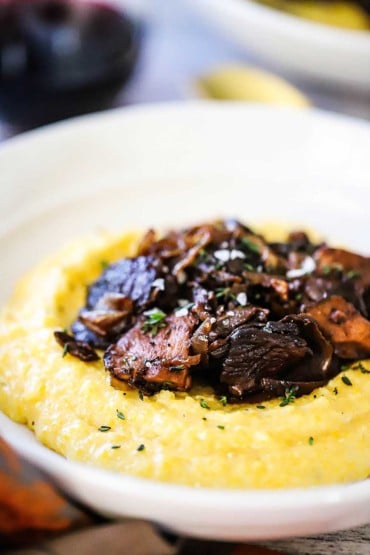 A close-up view of a white serving bowl filled with a mound of creamy polenta and topped with caramelized onions and porcini mushrooms.