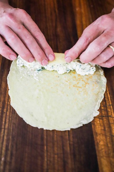 A person using his hands to roll a homemade crêpe filled with soft white cheese and herbs.