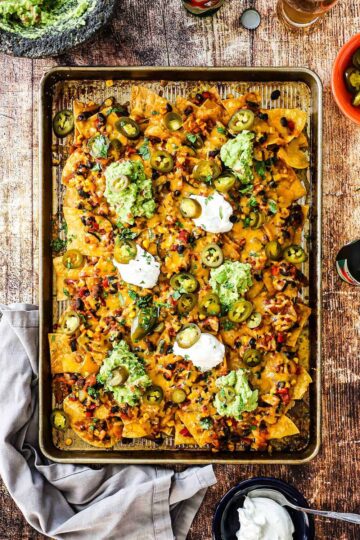 A large rectangular sheet pan piled high with nachos topped with dollops of guacamole and sour cream.