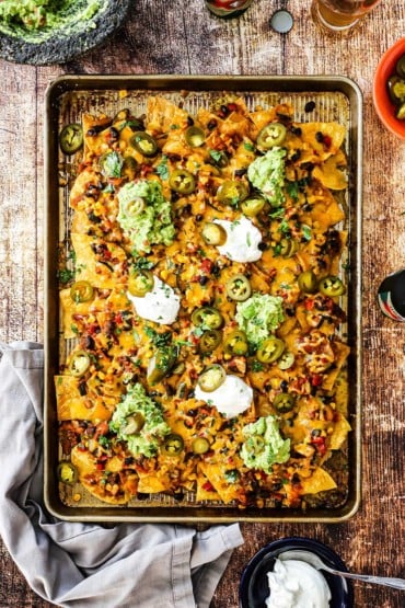 A large rectangular sheet pan piled high with nachos topped with dollops of guacamole and sour cream. 