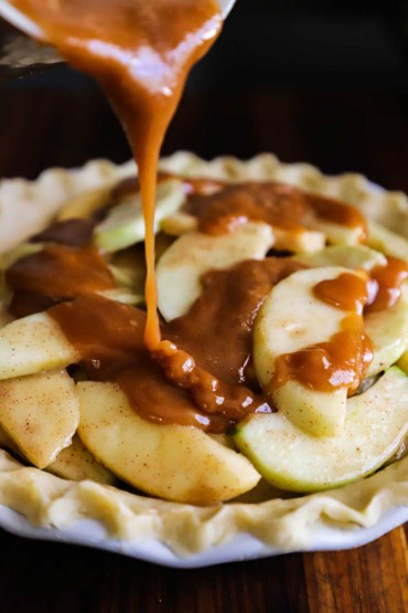 Caramel sauce being poured over a pie dish filled with sliced apples in a pie dough with crimped edges.
