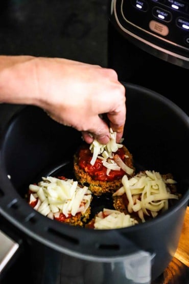 A person adding shredded mozzarella cheese to the top of breaded air-fried eggplant medallions in the basket of an air fryer.