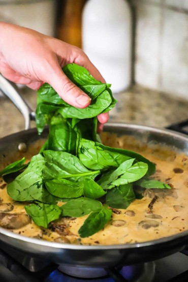 A person adding fresh spinach into a skillet filled with a creamy mushroom sauce.