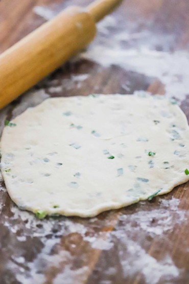 An uncooked scallion pancake sitting on a floured wooden cutting board next to a rolling pin.