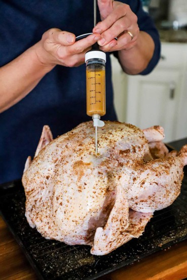 A person using a large meat syringe to inject a butter marinade into a seasoned raw turkey.
