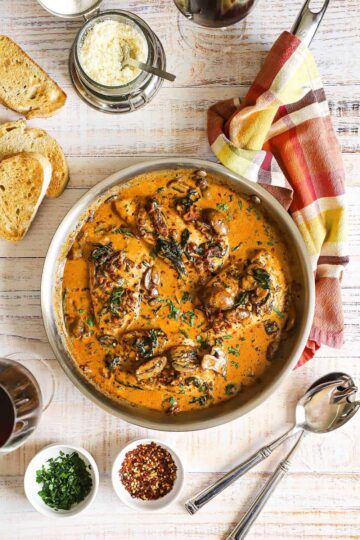 An overhead view of a large silver skillet filled with creamy Tuscan chicken with slices of toasted bread and a bowl of grated cheese nearby.
