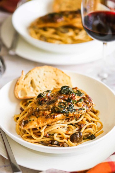 A white pasta bowl filled with spaghetti topped with creamy Tuscan chicken and a slice of toasted bread on the side.