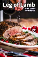 A person sprinkling coarse sea salt over the top of a sliced boneless leg of lamb with garlic and herbs on a platter.