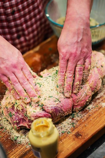 A pair of hands pressing an herb breadcrumb mixture into a leg of lamb that has Dijon mustard spread all over it.