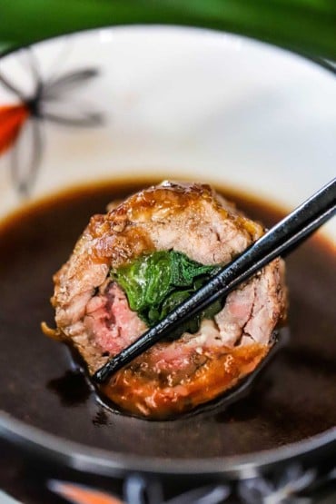 A slice of a thinly rolled piece of beef stuffed with scallions that is being held by chopsticks and lowering into a bowl of Asian sauce.