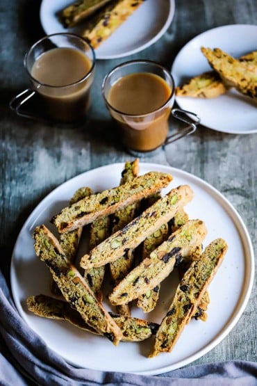 An overhead view of a white plate filled with a stack of biscotti with pistachios and dried cherries sitting next to two glass mugs of coffee.