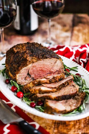 A close-up view of a roasted boneless leg of lamb with garlic and herbs sitting on white platter garnished with rosemary sprigs and cranberries.