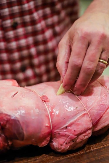 A person inserting garlic slivers into small slices that have been cut into an uncooked leg of lamb.