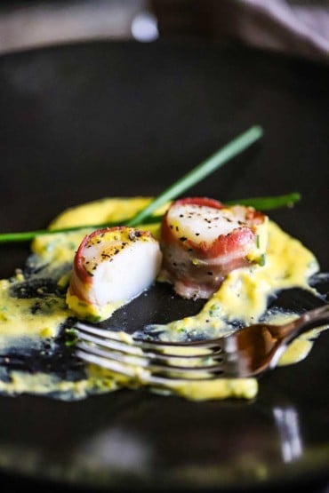 A pancetta-wrapped scallop that has been split open and is sitting in a yellow egg and wine sauce.