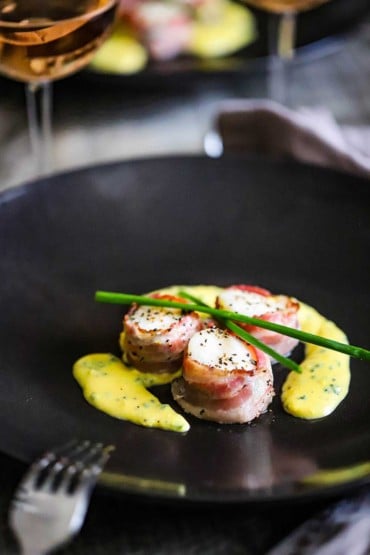 Three sea scallops with pancetta sitting on a black plate with a yellow egg and chive sauce on the plate, as well.