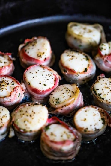 A black oval baking dish filled with sea scallops that have been baked with thin strips of pancetta wrapped around them.