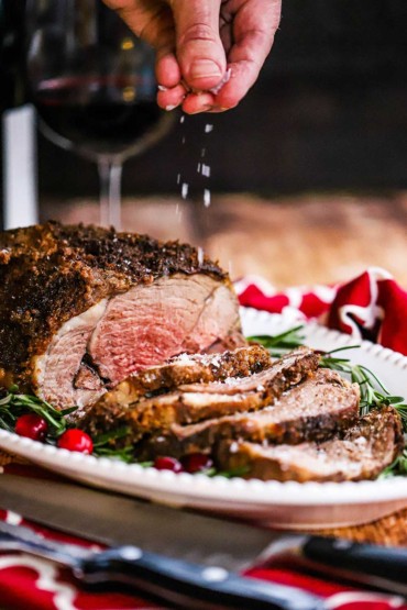 A person sprinkling coarse sea salt over the top of a sliced boneless leg of lamb with garlic and herbs on a platter.