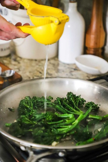 A person using a citrus press to transfer fresh lemon juice into a skillet filled with sautéd broccolini.