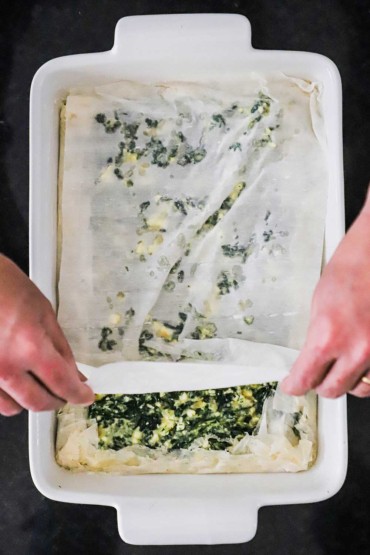 An overhead view of a person placing a thin sheet of phyllo dough in a rectangular baking dish that is filled with a spinach and cheese mixture.