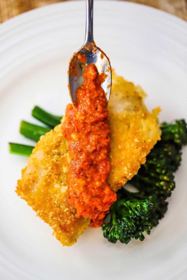 A small spoon being used to add romesco sauce over the top of Parmesan-crusted cod on top of a bunch of sautéed broccolini.