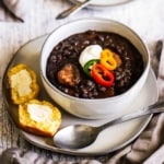 A close-up view of a soup bowl filled with black bean and andouille sausage topped with sliced peppers and a dollop of sour cream on a plate with a piece of cornbread on it.
