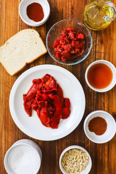 An overhead view of romesco sauce ingredients including roasted red peppers, almond slivers, roasted tomatoes, a slice of white bread, oil, and seasonings.