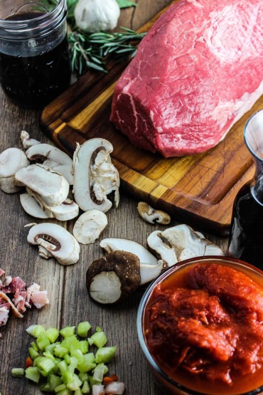 A cutting board holding an uncooked rump roast of beef and surrounded by sliced mushrooms, tomato sauce, vegetables, and beef stock.