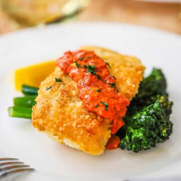 A white dinner plate filled with Parmesan crusted cod with romesco resting on sautéd broccolini.